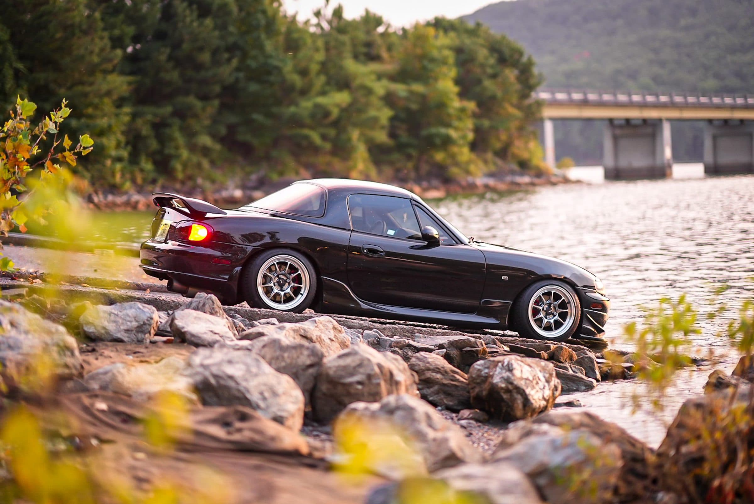 Stephen's black NB Miata with a rear wing on the rocks beside a lake